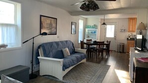 Ein Wohnzimmer mit blauem Sofa, einem Esstisch mit Stühlen, einem Deckenventilator und einer Küche im Hintergrund.
