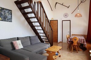 A living room with a grey sofa, a wooden table, a wooden chair, a staircase, and a dining table with a checkered tablecloth.