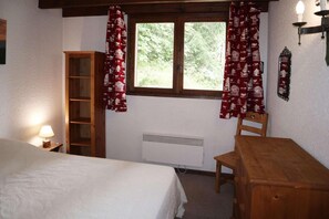 Une chambre à coucher avec un lit, une chaise en bois, un bureau, une armoire et une fenêtre avec des rideaux.