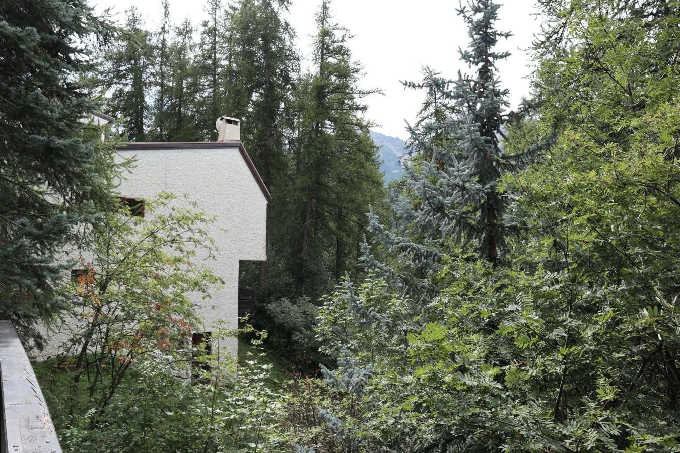 A white building with a sloped roof surrounded by dense forest and mountains in the background.