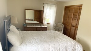 Un lit bien fait, recouvert d’une couette blanche, une table de chevet en bois avec une lampe, un miroir et une chaise en osier.