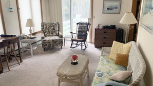 Un salon comprenant un canapé, deux fauteuils, une petite table et un fauteuil à bascule. Il y a une fenêtre avec des rideaux, une lampe et un tableau encadré au mur.
