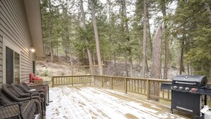 Une terrasse couverte équipée d’un barbecue, de meubles en osier et offrant une vue sur une zone boisée.