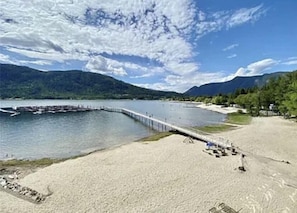 Uma praia à beira do lago com um píer de madeira que se estende na água, cercada por montanhas e árvores.