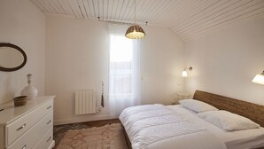Un dormitorio con cama, cómoda, lámpara colgante, radiador y ventana con cortinas traslúcidas.