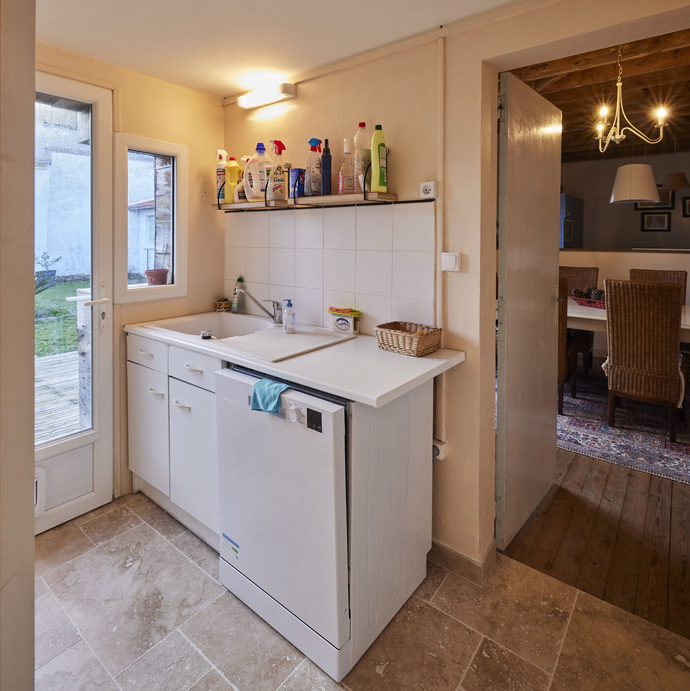 A compact kitchen with a built-in dishwasher, a window, and a shelf with cleaning supplies.