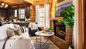Un salon chaleureux avec une cheminée, un canapé, une table à manger et un coin cuisine doté de meubles en bois et d’une grande fenêtre.