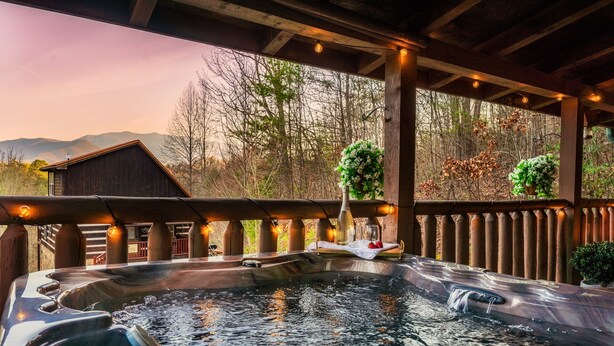 Un espace avec un bain à remous, une terrasse en bois, une vue sur les arbres et les montagnes, et un petit bâtiment en arrière-plan.