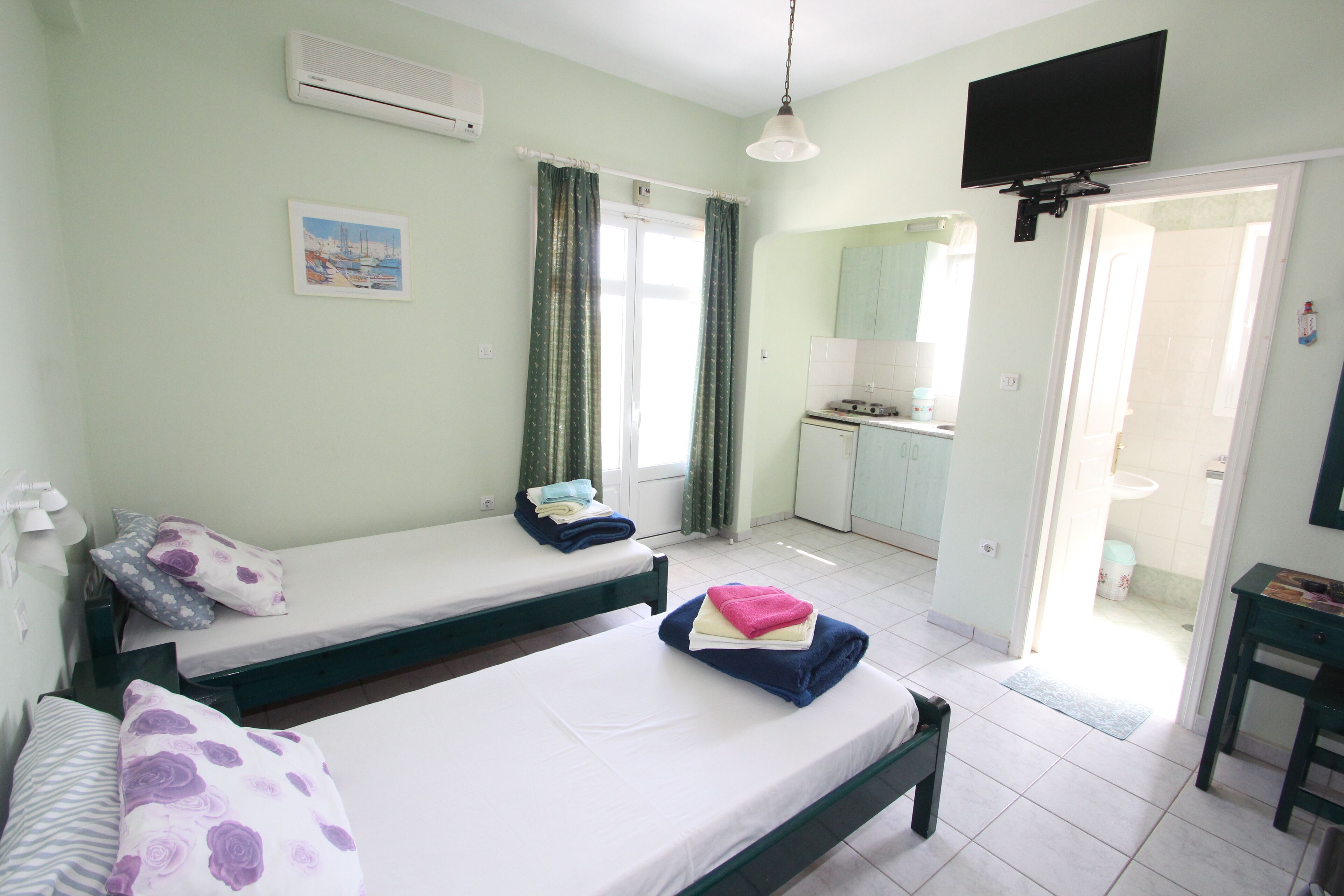 A room with two beds, a television, a kitchen area, and a bathroom visible through an open door.