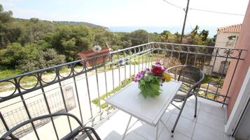 A balcony with a table and chairs, a flower arrangement, and a view of the sea and greenery.