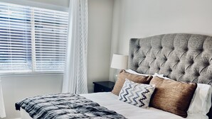 Quarto moderno com cabeceira estofada, cama grande com manta estampada, mesa de cabeceira com luminária e janela com persianas.