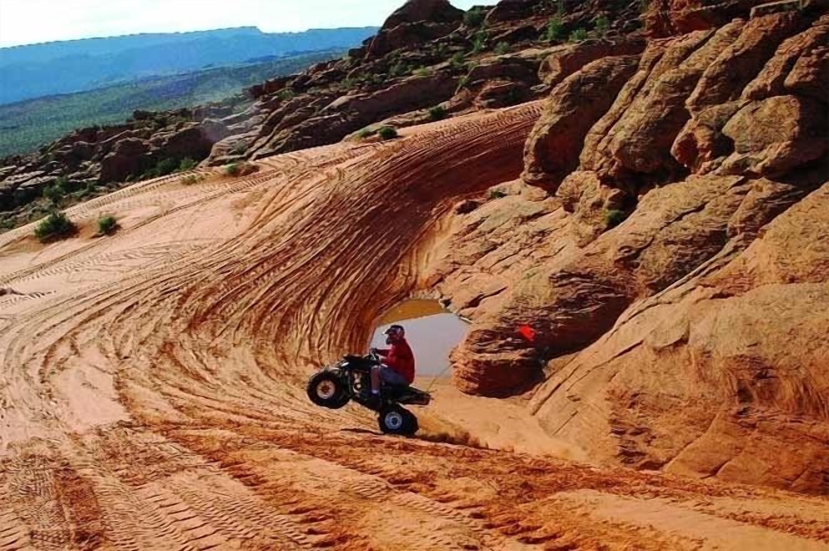 Una persona guida un quad attraverso un sentiero sterrato, con un paesaggio roccioso e un cielo sereno.