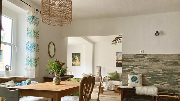 A spacious living room with a wooden dining table, comfortable seating, a fireplace, and a large window with curtains. - Gemütliche Ferienwohnung Ganz nah am Fluss (Roding)