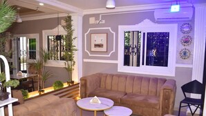 A cozy living area with a large beige sofa, two round tables, and a black chair. There are potted plants and a framed picture on the wall. -  Bulon Hotel (Yaoundé)