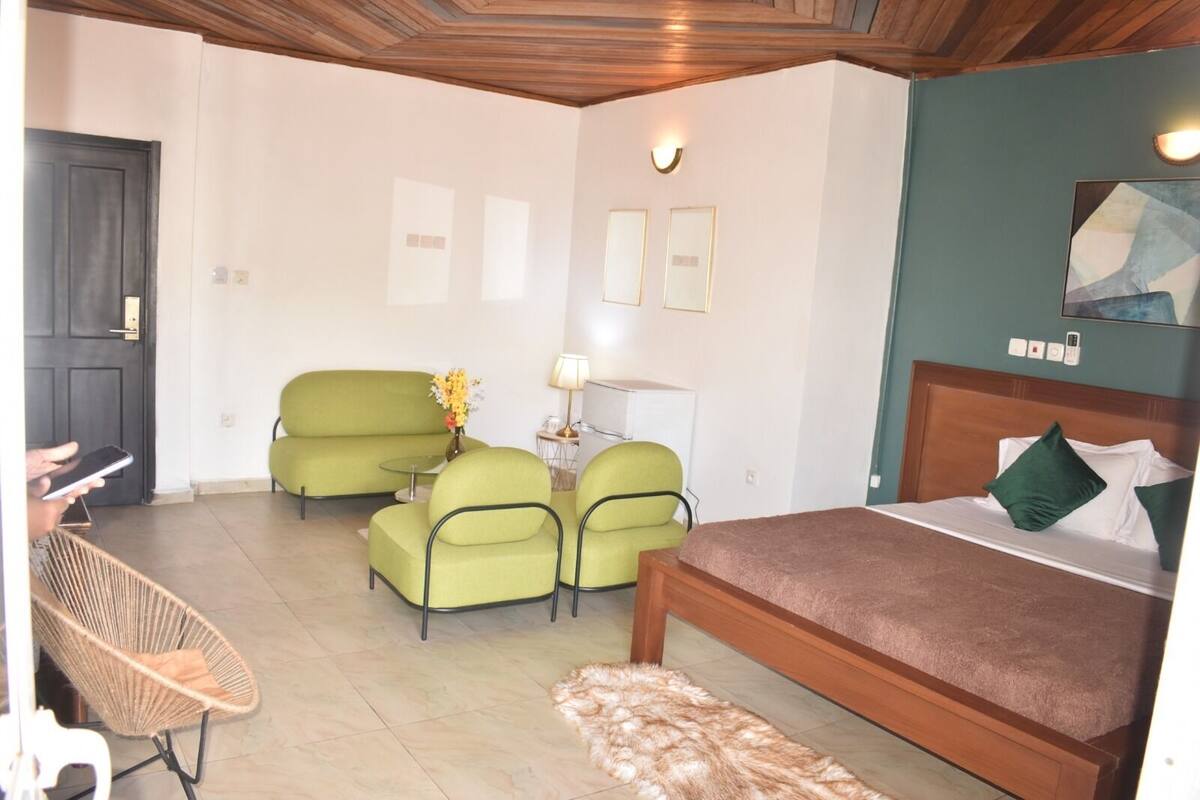 A hotel room with a bed, a green sofa, two armchairs, a small table with a lamp, and a wicker chair.