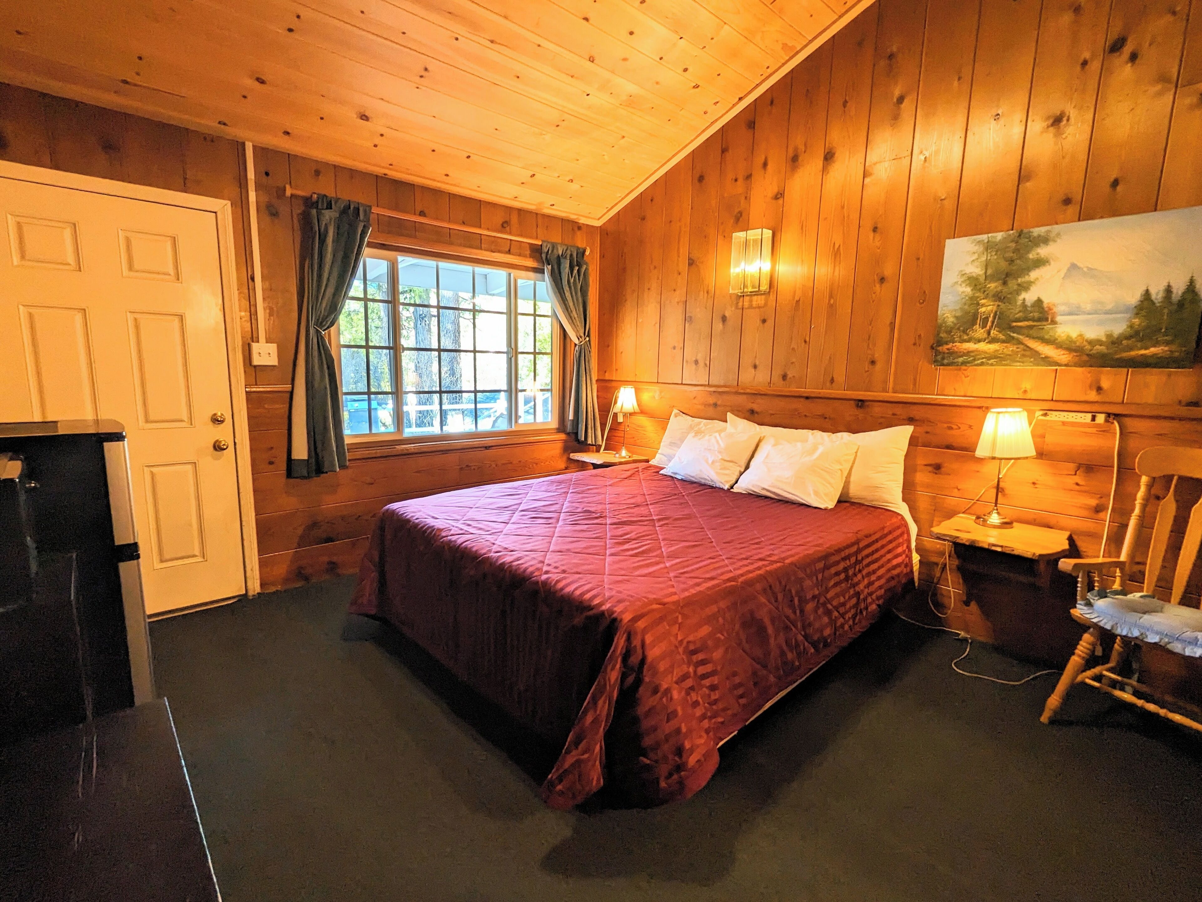 A wooden-paneled room with a bed, a painting, and a window with curtains.