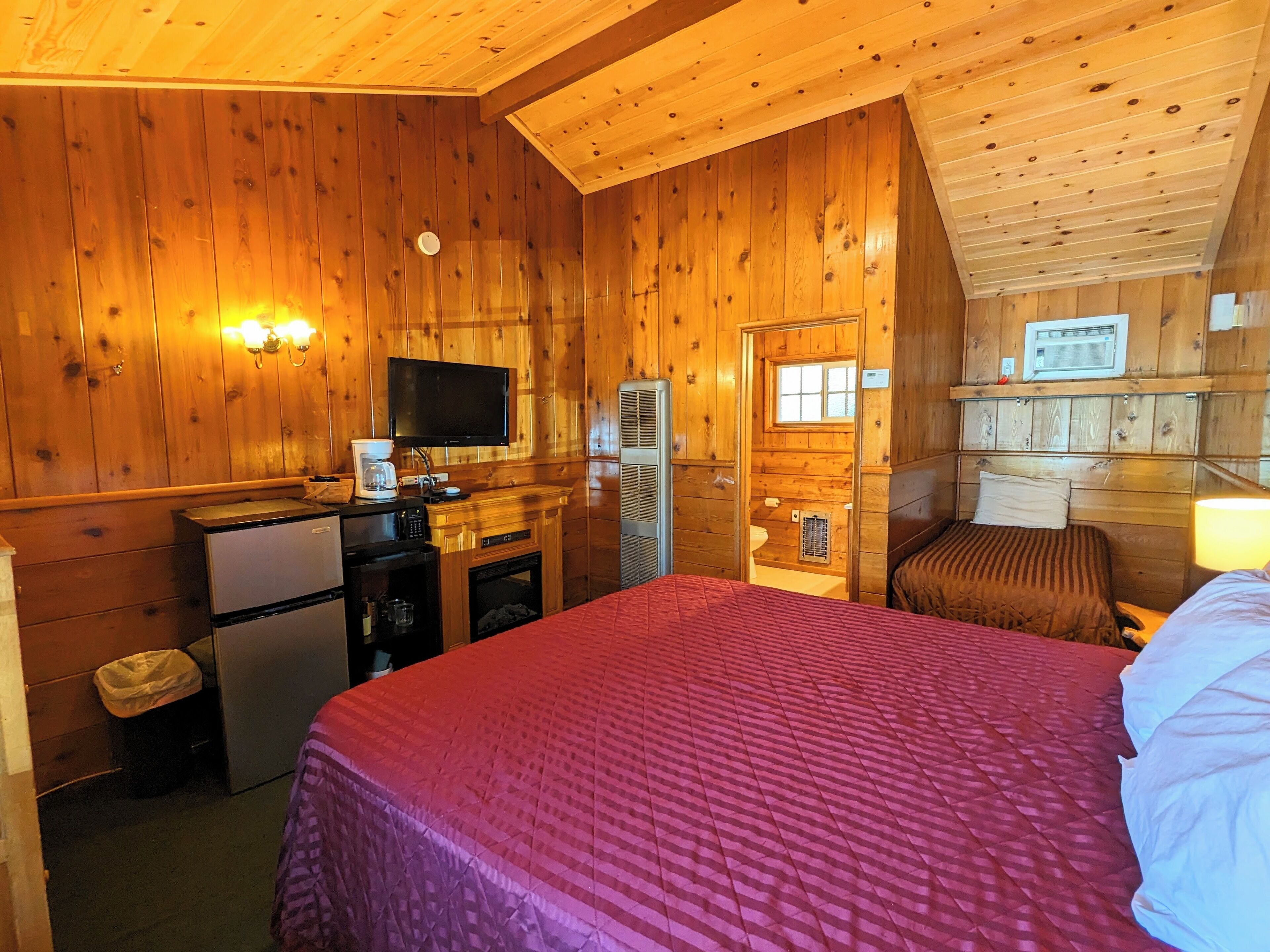 A cozy cabin-style room with a bed, a small kitchenette, a television, and a window.