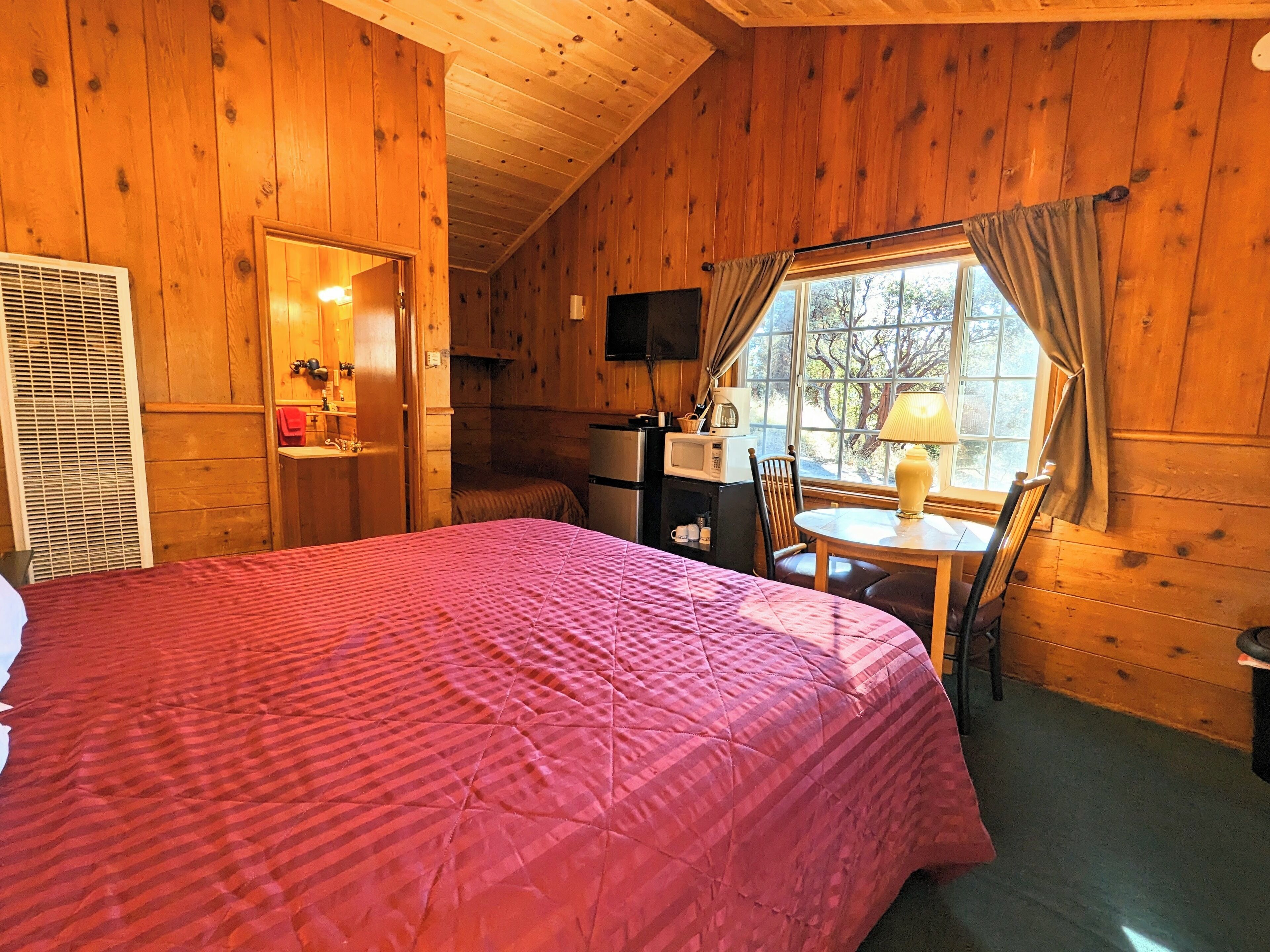 A cozy cabin room with a bed, a small kitchenette, a dining table with chairs, and a window with curtains.