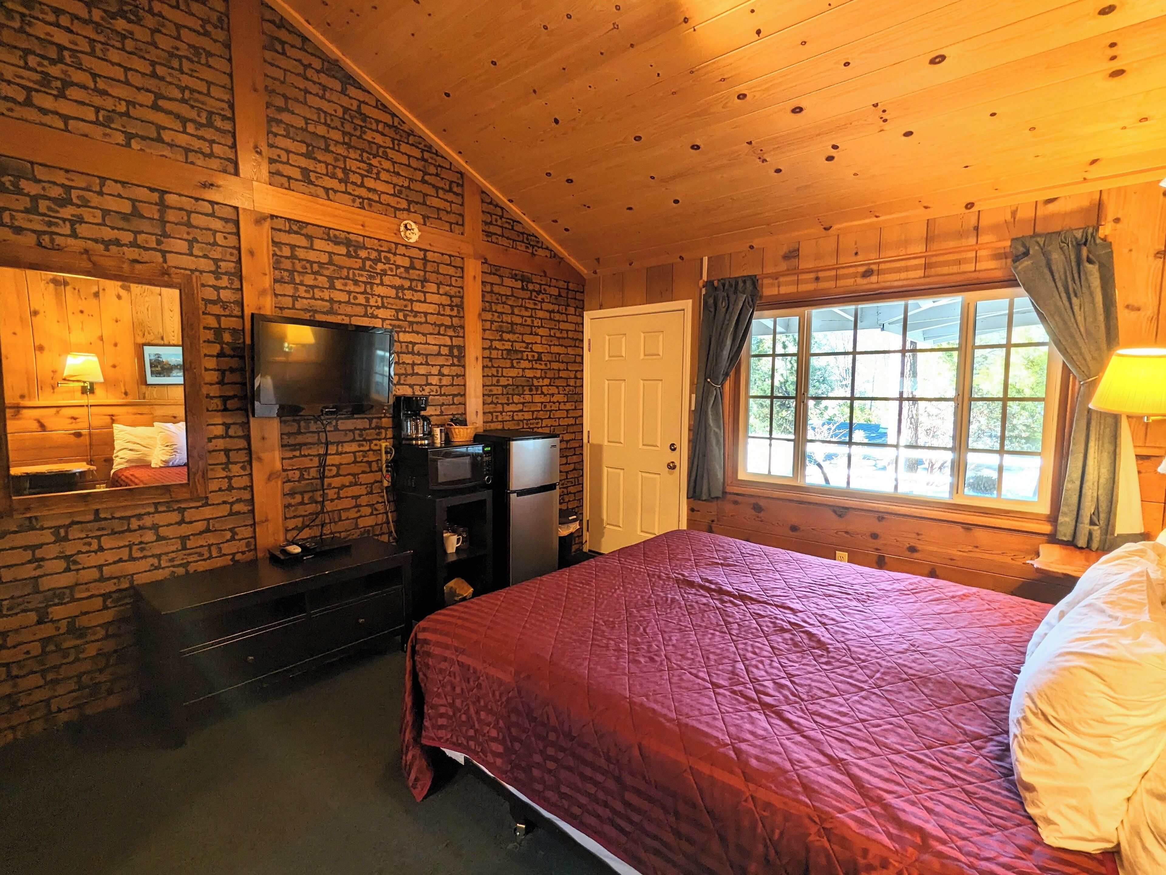 Ein gemütliches Zimmer mit Backsteinwand, Holzbalkendecke, einem Bett mit roter Bettdecke, einem Fernseher, einer kleinen Küchenzeile und einem Fenster mit Vorhängen.