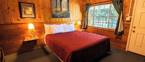A wooden room with a bed, two bedside lamps, a window with curtains, and a door.