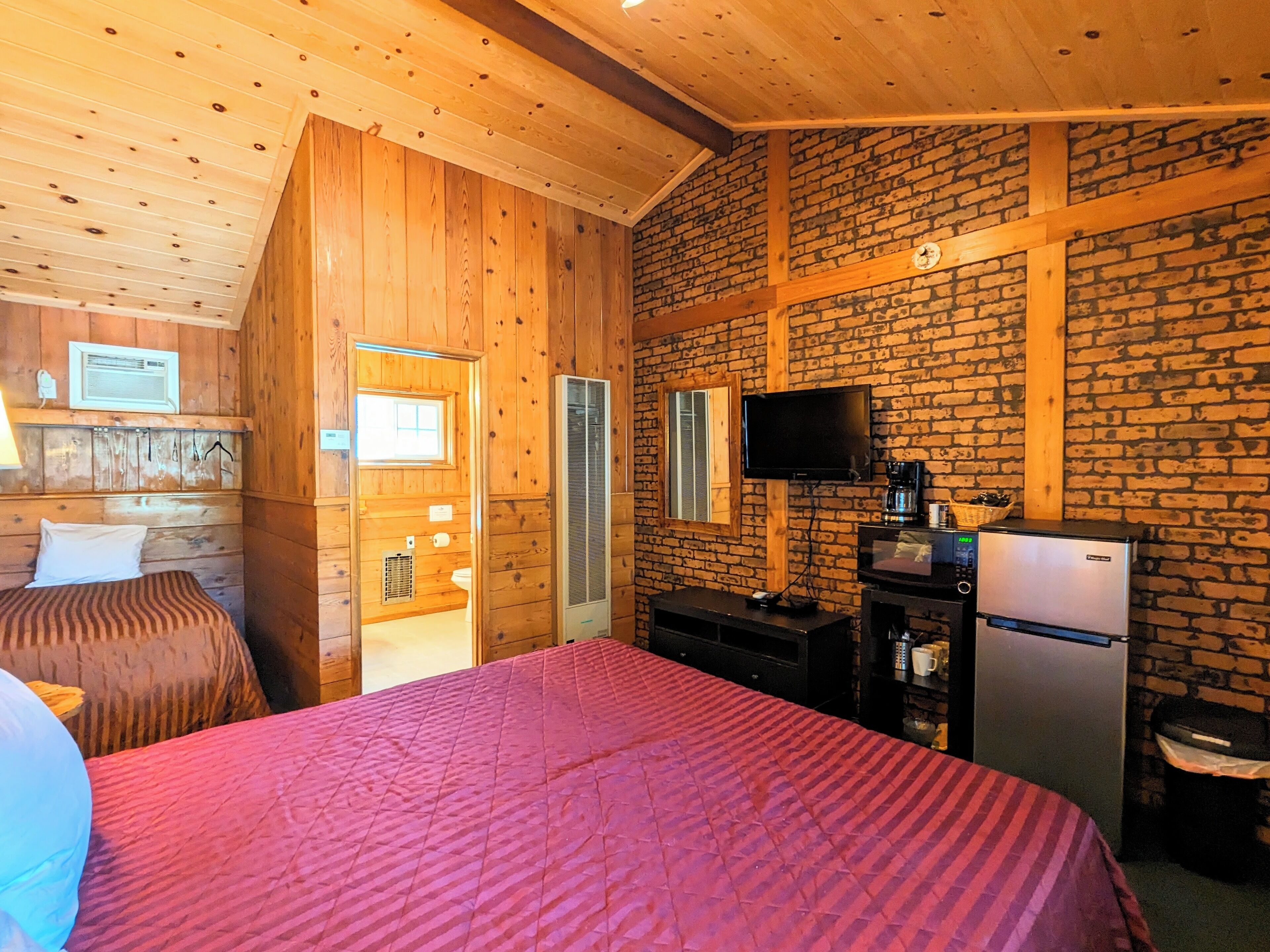 Ein Zimmer in einer Holzhütte mit einem Bett, einem Fernseher, einem Kühlschrank und einer Ziegelwand.