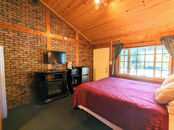 A cozy room with a brick wall, wooden ceiling, a bed with a red blanket, a TV, a coffee maker, and a refrigerator. - Bluebird Cottage Inn (Idyllwild)