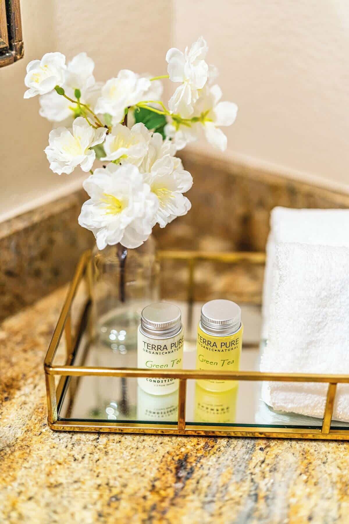 A bathroom vanity with a glass tray holding two bottles of Terra Pure organic-natured shampoo and conditioner, a white towel, and a vase with white flowers.