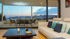 Una sala de estar moderna con un gran ventanal que da a un balcón exterior, un sofá blanco con cojines, una mesa de centro con botellas y aperitivos, y vista a la ciudad al atardecer.