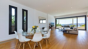 Un moderno salón con una mesa de comedor de cristal, sillas blancas, un sofá y un amplio ventanal con vistas a la ciudad.