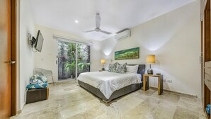 Ein Schlafzimmer mit einem Bett, zwei Nachttischen, einem Deckenventilator, einer Klimaanlage, einem Fenster mit Blick auf Grünflächen und einer Tür.