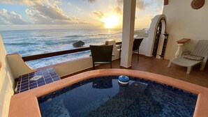 Un balcone con vista sull'oceano al tramonto, una vasca idromassaggio e un'area relax con sedie e un tavolo.