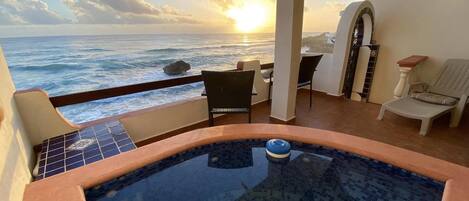 A balcony with a view of the ocean at sunset, a hot tub, and seating area with chairs and a table.