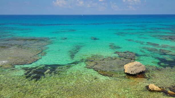 Vista limpida su un mare turchese, con rocce visibili sott'acqua e una barca in lontananza.