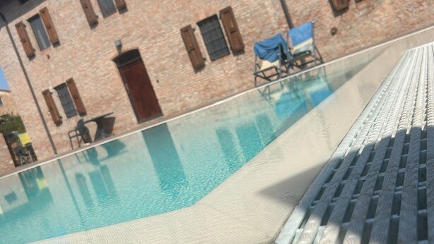 Ein Schwimmbecken mit gefliestem Sonnendeck, ein Backsteinhaus mit Fenstern und Fensterläden sowie ein Stuhl am Pool.