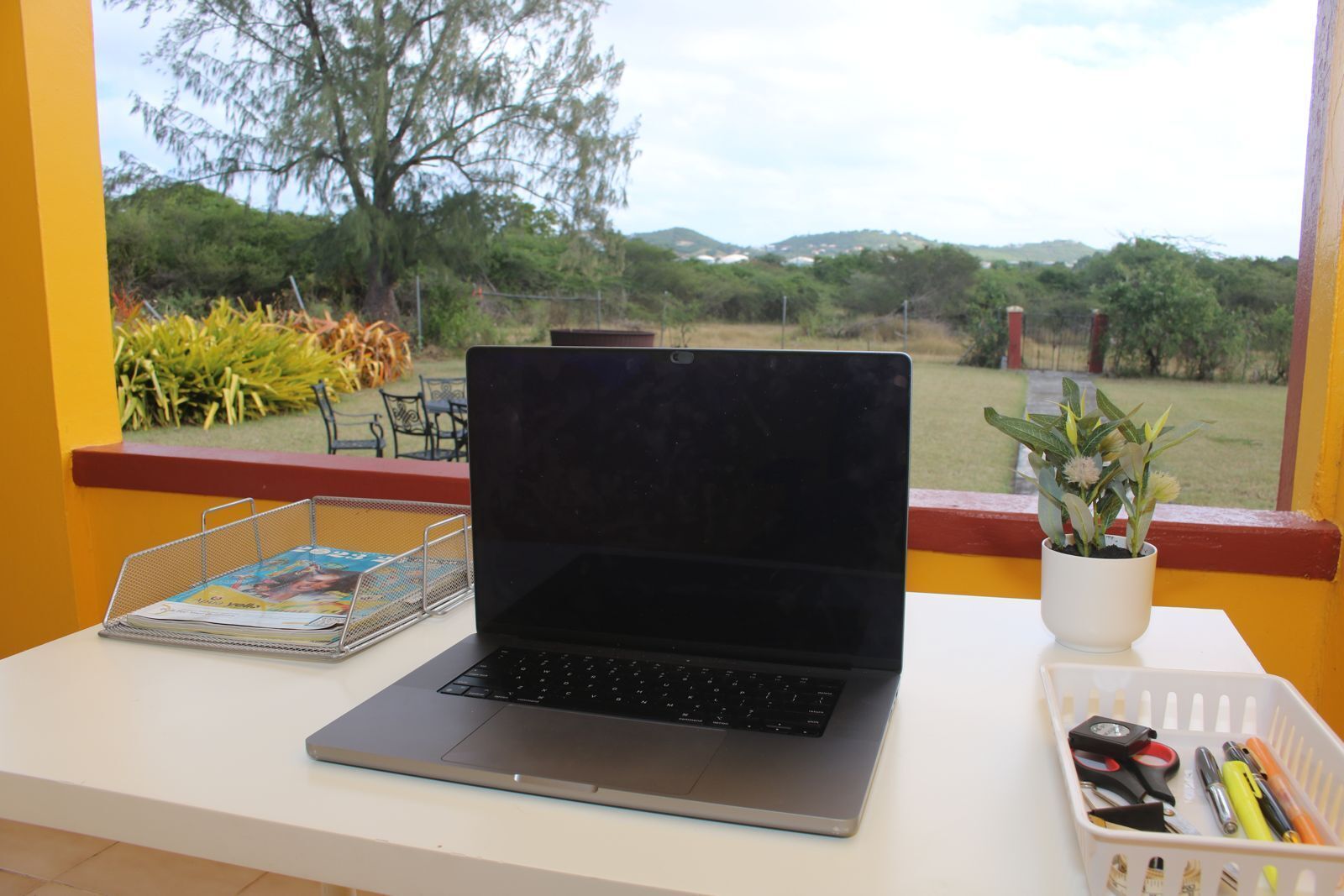 Ein Laptop auf einem Tisch mit einer Zeitschrift, einer Pflanze und einem Stiftekörbchen.