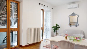 Una habitación con puerta de madera, cortinas blancas, radiador, espejo, mesa con sillas y una planta.