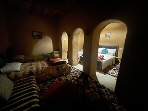 A dimly lit room with a bed, a small table, and a chair. There are arched doorways leading to another room. - Maroc des Merveilles - Chez l'habitant (Ait Sedrate Sahl El Gharbia)