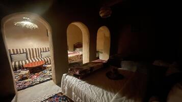 A dimly lit room with a sofa, a bed, and a small table. There are two arches in the room, and a colorful rug is visible on the floor.