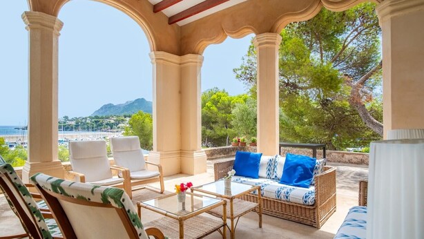 A covered outdoor seating area with chairs and a sofa, overlooking a scenic view of the sea and hills.