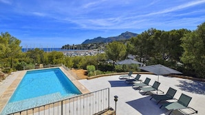 A swimming pool with lounge chairs and umbrellas, surrounded by greenery and a view of the sea and hills.