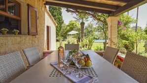 Un espace extérieur abrité, aménagé pour les repas, avec des meubles en osier, une table dressée avec des verres à vin et des fruits, et une vue sur un jardin avec des arbres et un bâtiment en arrière-plan.