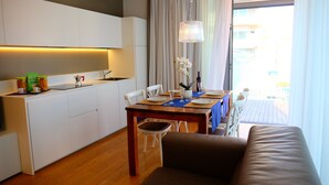 A modern kitchen with white cabinetry, a dining area with a wooden table and chairs, and a view of the outdoors through large windows. - Apartment 'Residence Shine 06' with Private Terrace and Air Conditioning (Jesolo)
