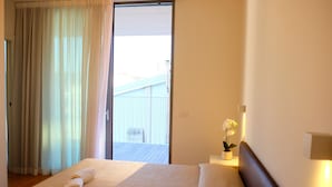 A hotel room with a bed, a window with curtains, a small table with a lamp, and a potted plant. - Apartment 'Residence Shine 06' with Private Terrace and Air Conditioning (Jesolo)