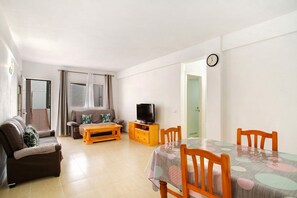 Ein Wohnzimmer mit einer Couch, einem Fernseher auf einem Holzboden, einem Esstisch mit Stühlen und einer Wanduhr.