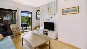 Una sala de estar moderna con televisor, una mesa de centro de madera, un sofá y una silla. Una escalera conduce a un balcón con vistas a zonas verdes.
