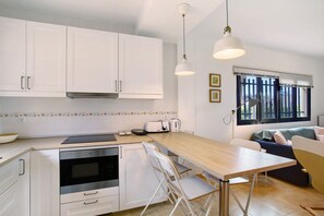 Cozinha moderna com armários brancos, forno embutido, área de jantar com mesa e cadeiras, e janela com persianas.