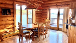 A wooden cabin interior with a dining table, chairs, and a view of a lake through large windows.