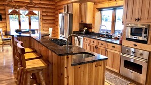 A log cabin kitchen with a central island, stainless steel appliances, wooden cabinets, and a large window.