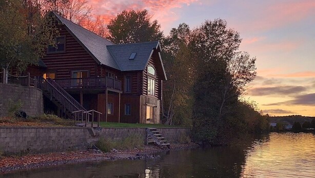 A log cabin with a balcony, a staircase leading to the entrance, and a river in the foreground.