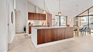 Une cuisine moderne avec des meubles en bois, un îlot central, des luminaires suspendus et de grandes fenêtres offrant une vue sur l’extérieur.